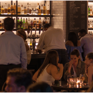 Lively bar scene with patrons seated and standing around a well-stocked counter, bartenders serving drinks, and dim lighting creating a warm ambiance—used to illustrate AML training context for hospitality venues.